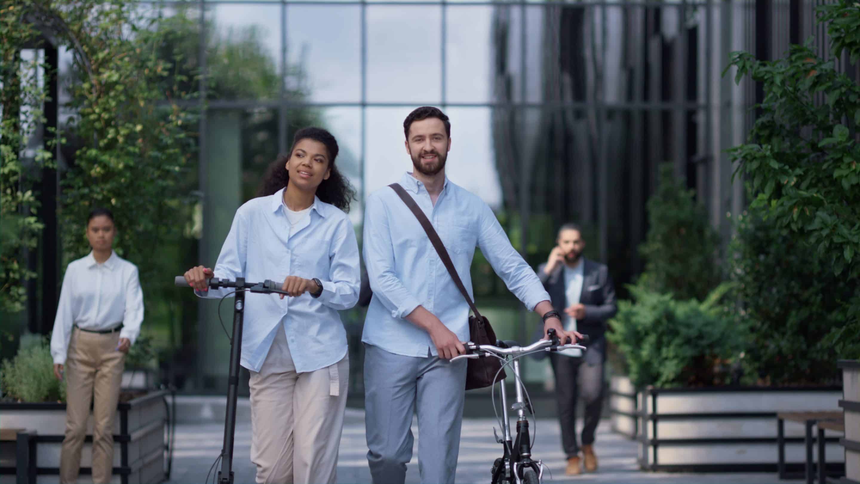 La photo montre deux jeunes adultes, un homme et une femme, marchant côte à côte dans un environnement urbain moderne et verdoyant. La femme pousse une trottinette électrique tandis que l'homme marche avec un vélo pliant. Tous deux sont habillés de manière décontractée et professionnelle, portant des chemises bleu clair. En arrière-plan, on peut voir d'autres personnes se déplaçant, une femme marchant et un homme au téléphone, dans un environnement qui semble être un complexe de bureaux entouré de végétation. La scène dégage une impression de mobilité durable et d'équilibre entre vie professionnelle et bien-être personnel.