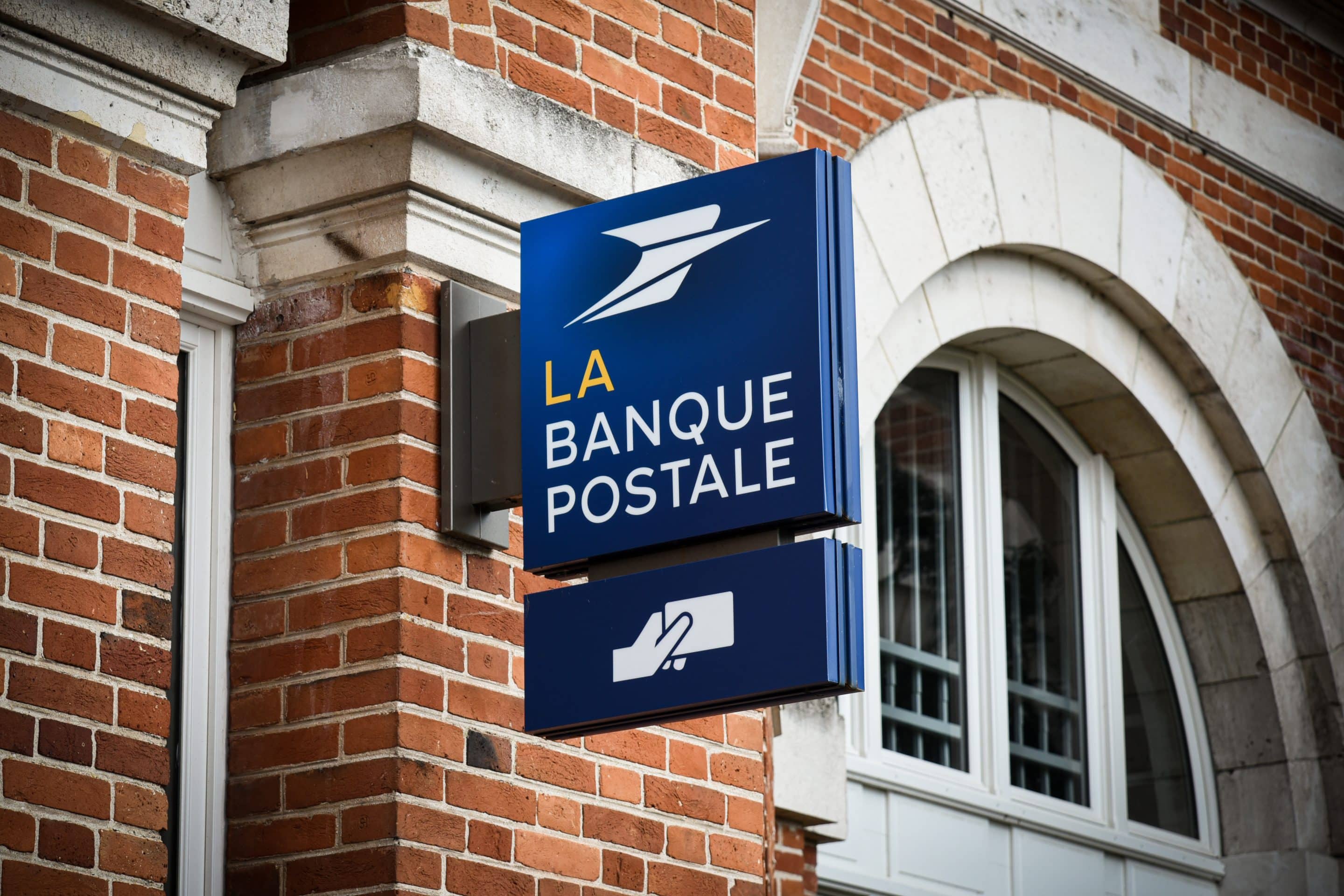 Photo d'une enseigne bleue de La Banque Postale, fixée sur la façade d'un bâtiment en briques rouges. L'enseigne affiche le logo de l'institution ainsi que le nom "LA BANQUE POSTALE" en lettres blanches et jaunes. Au-dessous, un pictogramme blanc représentant une main glissant un document, probablement une carte bancaire, est visible.